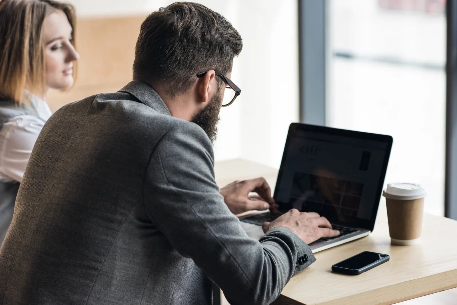 A person using a laptop while a colleague looks on beside a window
